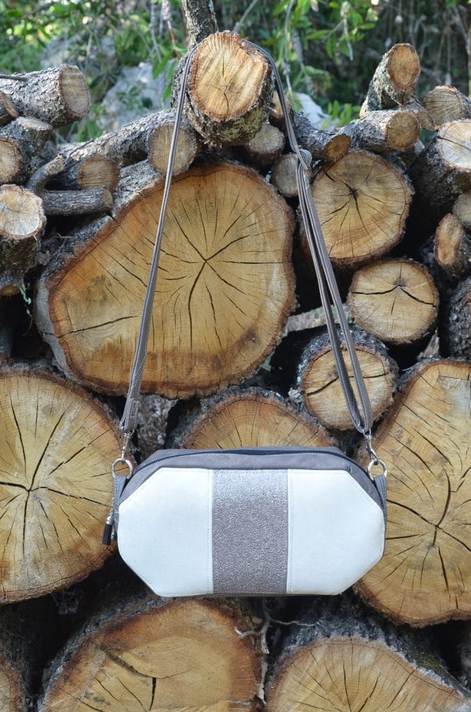 Petit sac en bandoulière blanc et gris avec une fermeture éclaire sur le dessus, suspendu à une bûche dépassant du tas de bûches.