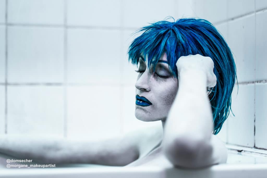 Bilal, Femme en robe de soirée noire décolletée aux épaules, debout dans une douche et une salle de bain délabrée, avec des cheveux bleus et une plaies sur la main droite, Body painting de l'artiste Morgane MakeUp artiste