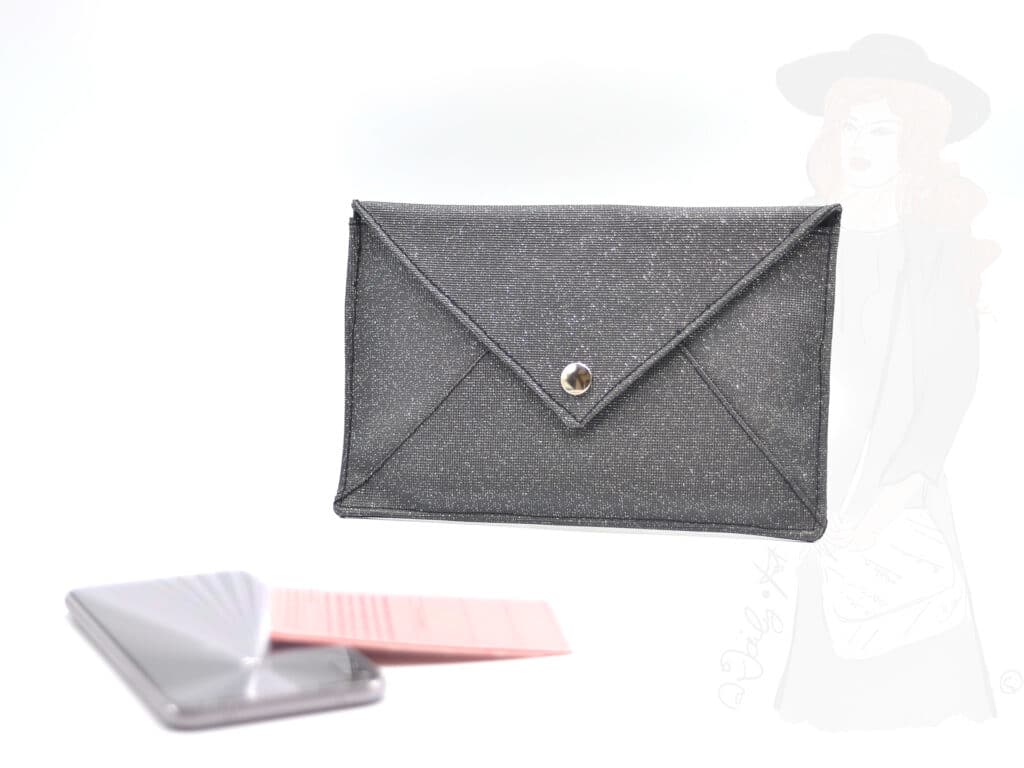 Pochette enveloppe noire argentée, vue de 3/4 avec un mobile et un permis rose posés à côté.