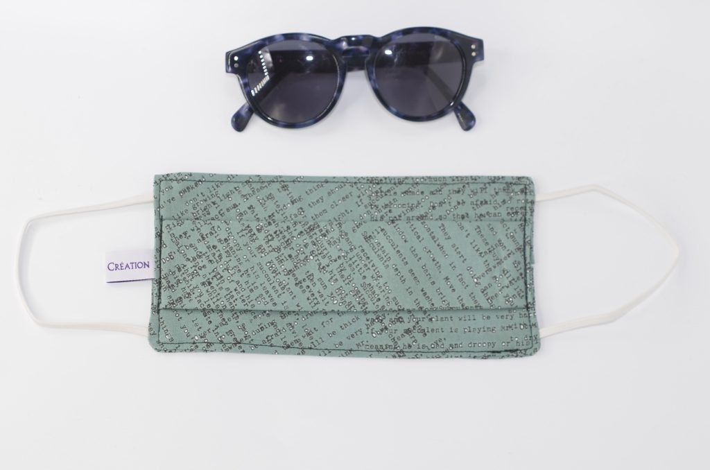 Masque en tissu vert avec des inscription noir; Lunettes de soleil posée sur un fond blanc
