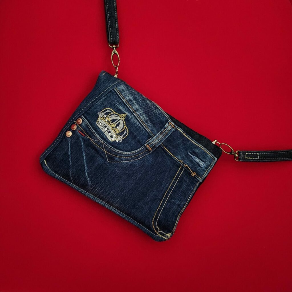 Pochette enveloppe en jean de récupération, avec une bandoulière. Pochette vue de dos, posée sur du velours rouge, tout ce qu'il y a de plus élégant pour une femme.