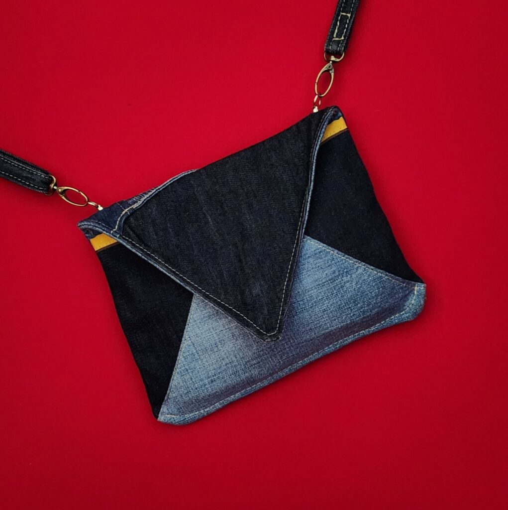 Pochette enveloppe en jean de récupération, avec une bandoulière. Pochette posée sur du velours rouge, tout ce qu'il y a de plus élégant pour une femme.